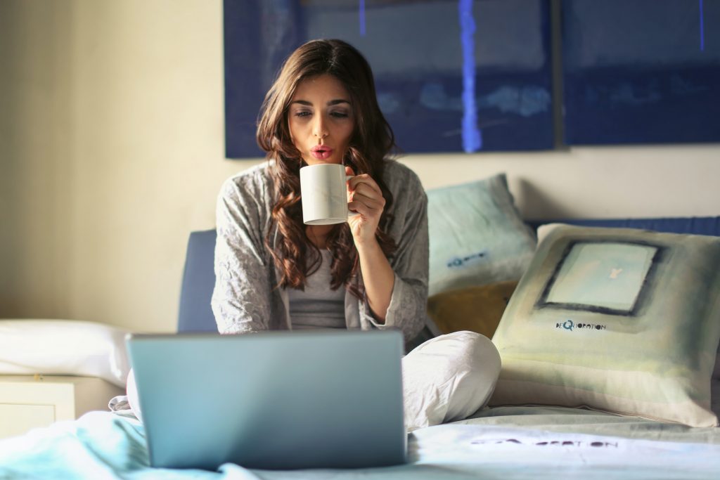 woman-in-grey-jacket-sits-on-bed-uses-grey-laptop