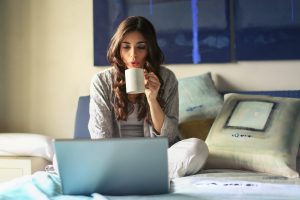 woman-in-grey-jacket-sits-on-bed-uses-grey-laptop